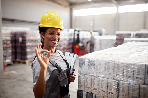 Happy African American female worker showing ok sign while working in supply chain and looking at camera.