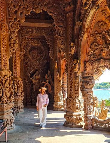 A woman in a hat is casually strolling through a labyrinthine building, his silhouette framed against the intricate play of light and shadows. Sanctuary of Truth Pattaya