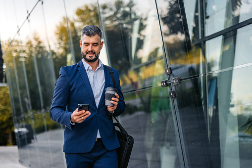 Handsome mid-adult man using his smart phone while drinking coffee in front of an office building.