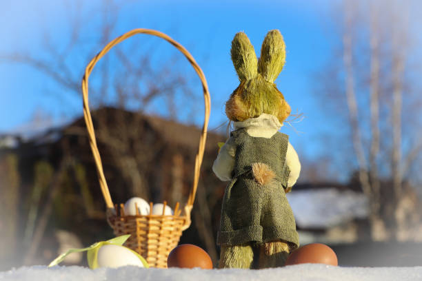A toy Easter bunny stands with its back turned. Blue sky background. Nearby is a decorative basket with eggs.