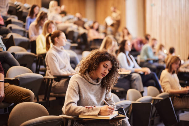 Writing an exam at the university! Female college student writing an exam during a class at lecture hall. Her classmate are in the background. education stage stock pictures, royalty-free photos & images