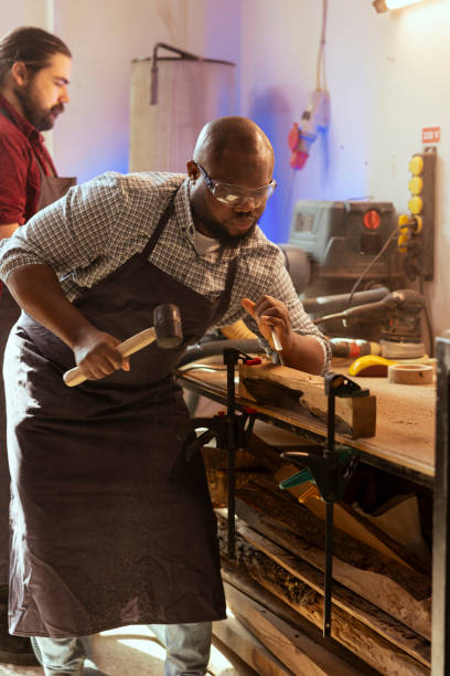 istock Craftsperson carving designs into wood using chisel and hammer next to coworker 2122194466