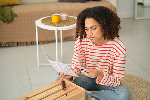 Elevated view of an african young Woman reading manual to assembly new furniture at home