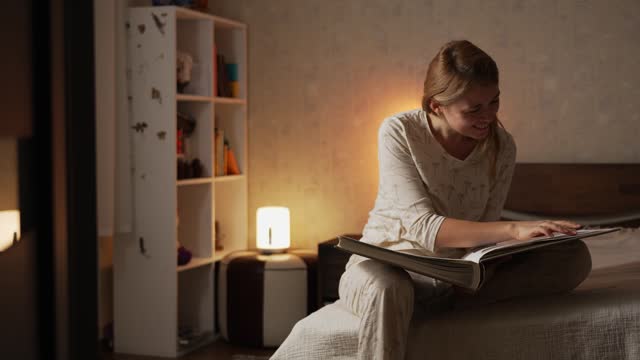 Happy smiling blonde woman in pajamas sitting on bed in cozy comfortable bedroom watching a photo album with photographs