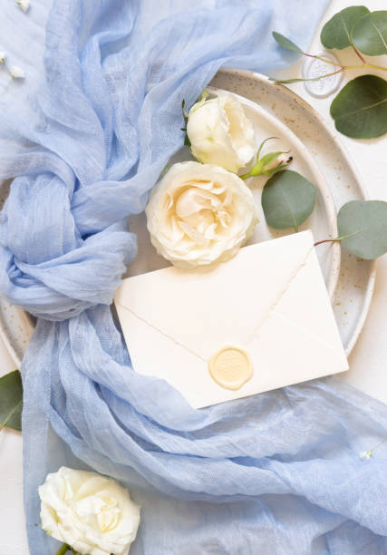 Envelope near light blue fabric knot and cream roses on plates top view copy space, wedding mockup