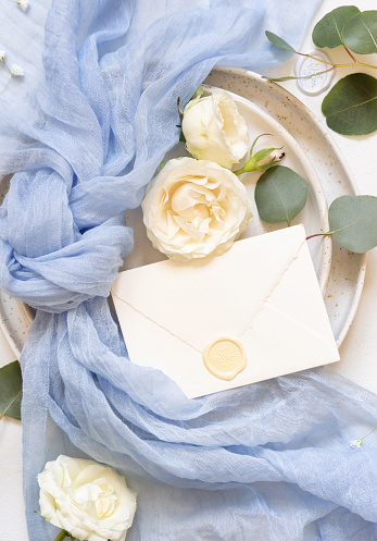 Sealed envelope near light blue fabric knot and cream roses on plates top view, copy space. Wedding stationery mockup. Romantic table place with sealed envelope, flowers and pastel decor