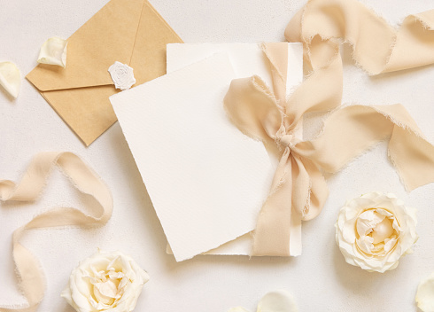 Cards tied with a beige silk ribbonnear cream roses and sealed envelope on white table top view, mockup. Romantic scene with vertical blank cards. Wedding, Valentines, Spring or Mothers day stationery
