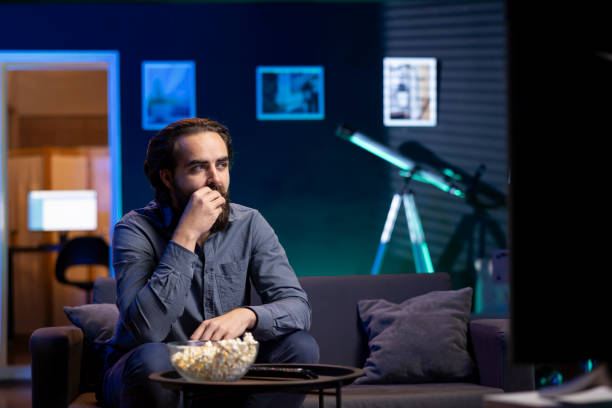 istock Man eating popcorn on couch, watching news broadcast on TV 2122229410