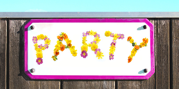 The word PARTY made of yellow pink red and orange flowers collage, enamel, metal or pottery sign on a rustic wooden wall. pink frame, under a bright turquoise sky. the sun is shining, elongated stone slab with copy space to write in retro style for cards. fastened with four screws. floral lettering can be used as posters for weddings, anniversaries, corporate events, welcome parties and celebrations. real blossoms photo-collage