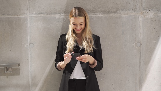 A young woman in a business suit is standing outdoors, using her smartphone to send a message.