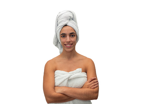 young woman, with a towel wrapped around her chest and head, stands with crossed arms, looking at the camera with a smile after a shower, radiating freshness and confidence on white background