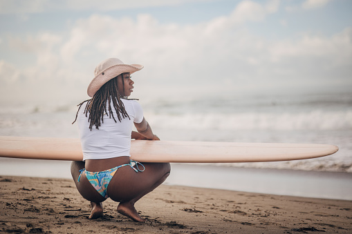 Capture the essence of tranquility and anticipation with this evocative image. A black woman squats gracefully on the sandy shore, surfboard in hand, gazing out at the rolling waves. The individual’s attire and poised stance embody the spirit of surfing, blending the thrill of the sport with moments of peaceful reflection.