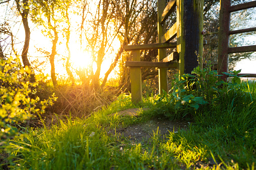 Springtime sun seen setting in a lush meadow next to a wooden steeple used as part of a public footpath in a rural area.