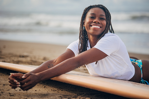 Black woman is captured in a serene moment, laying on a surfboard close to the shore. Woman appears to be soaking in the tranquil atmosphere, with the gentle waves and scenic horizon in the background offering a picturesque setting.
