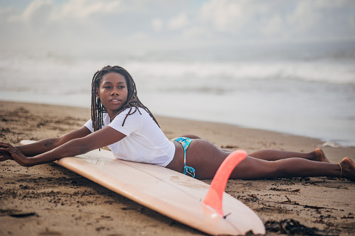 Black woman is captured in a serene moment, laying on a surfboard close to the shore. Woman appears to be soaking in the tranquil atmosphere, with the gentle waves and scenic horizon in the background offering a picturesque setting.
