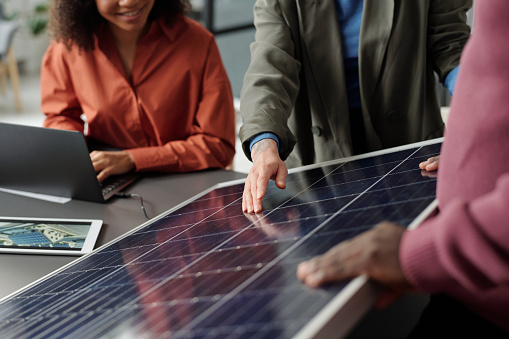 Cropped shot of young businesswoman in formalwear pointing at solar panel model while making presentation to colleagues by workplace