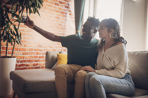 This image captures a warm and intimate moment between two individuals as they sit closely on a comfortable sofa, engrossed in sharing content on a smartphone. The cozy ambiance, accentuated by the soft lighting and the rustic charm of exposed brick walls, evokes a sense of connection and shared experiences.