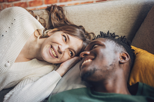 Lying on a cozy couch, a young multiracial couple shares an intimate moment filled with laughter and genuine connection. The woman leans in, her gaze affectionately locked with her partner's, as they revel in the joy of each other's company