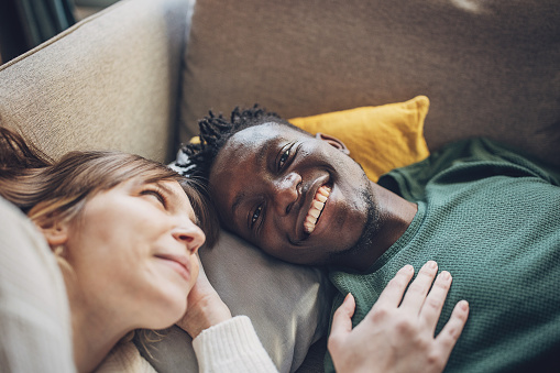 Lying on a cozy couch, a young multiracial couple shares an intimate moment filled with laughter and genuine connection. The woman leans in, her gaze affectionately locked with her partner's, as they revel in the joy of each other's company