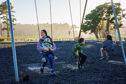 A group of brothers and sisters of Eurasian descent and ranging in age from 1 to 16 have fun swinging together at a neighborhood park on a sunny and cool day in Oregon.