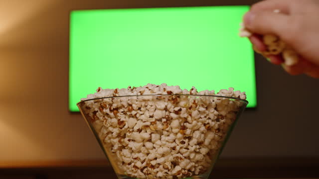 Two Hands of a Man and a Woman Simultaneously Take Popcorn from a Bowl in Front of the TV with a Layout of an Empty Green Screen.