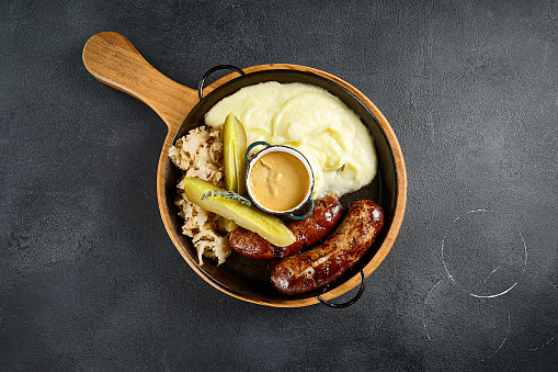 Top view of rustic grilled sausages with creamy mashed potatoes and tangy pickles, on a wooden server against a textured backdrop. Comfort cuisine at its finest.