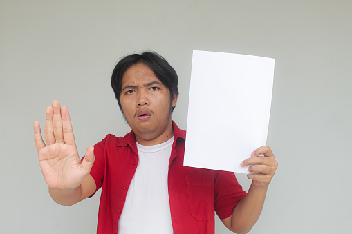 Close up portrait of young Asian man showing blank white paper with prohibiting gesture or stop gesture.