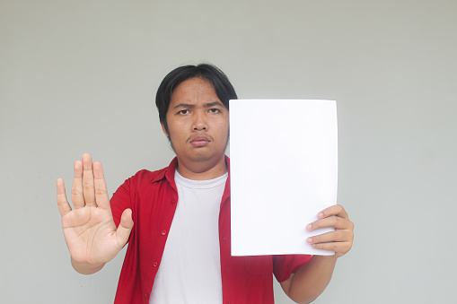 Close up portrait of young Asian man showing blank white paper with prohibiting gesture or stop gesture.