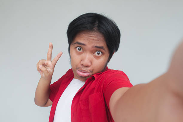 Young Asian man in red shirt taking selfie with funny expression, Isolated in gray background.