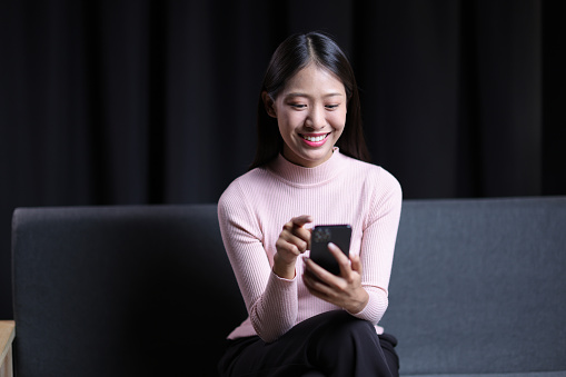 Young Asian woman sitting on sofa at home using mobile phone to send text messages and talk on the phone with her friends.