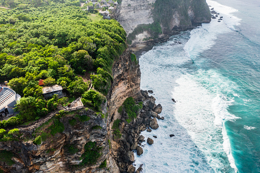 Lush greenery covers a steep cliff that descends to a rocky shoreline where turquoise ocean waves crash against the rocks, an aerial perspective capturing the dramatic contrast between land and sea. Bali, Uluwatu.