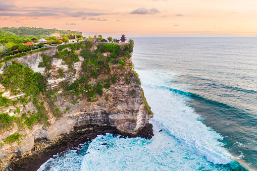 A lush green cliff, framed by crashing waves, stands tall against the backdrop of a vivid sunset. Uluwatu Temple (Pura Luhur Uluwatu) perched at the edge, overlook the vast expanse of the ocean.