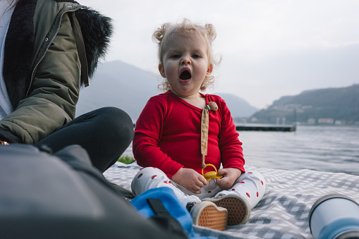 Toddler sits by mother and yawns by lakeshore during afternoon picnic