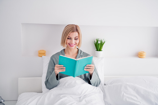 Photo portrait of blonde attractive young woman sit bed read book wear trendy gray pajama light bedroom interior minimalist design.