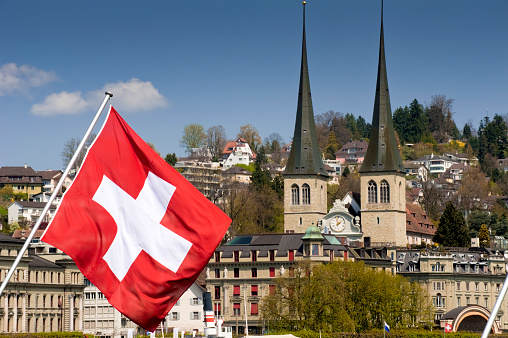 Raised Swiss national flag in front of the two towers of St. Leodegar's Church and hotels in Lucerne, Switzerland