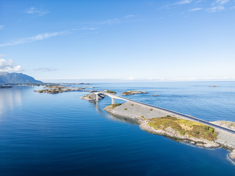 A modern bridge curving over calm blue ocean waters, linking small islands together. The bridge looks like it's floating on the water. The bottom of the sea can be seen near the islands, and big mountains in the far background add to the calm scene.
