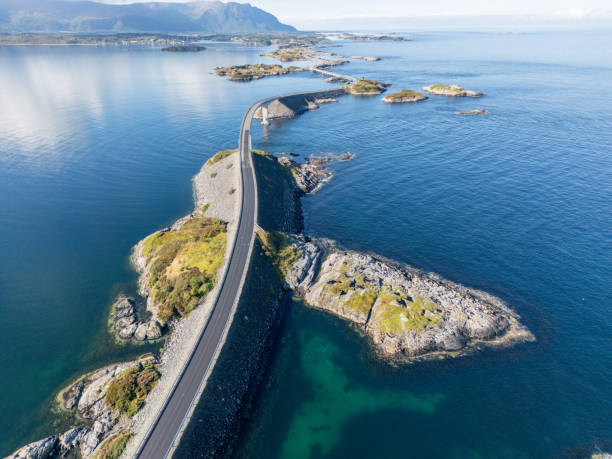 View of a road winding through rocky islands and skerries, under a clear sky. The road looks like a thin line connecting these small land pieces, made more beautiful by the calm sea around it. Cars on the road are tiny against the big landscape with clear skies and mountains in the far background