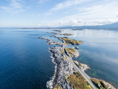 A narrow asphalt road curves through the rocky Norwegian coast, passing a series of small islands and islets. Its winding path provides a unique driving experience, set against the large, clear ocean and under skies reflected in the water.