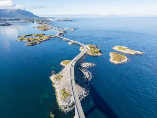 View of a long bridge over rocky islands and skerries, under a clear sky. The road looks like a thin line connecting these small land pieces, made more beautiful by the calm sea around it. Cars on the road are tiny against the big landscape.