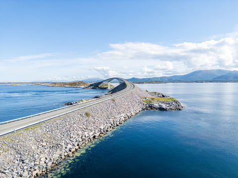 Side view of a  modern bridge stretching over calm blue waters, linking an island with the mainland. The design of the bridge stands out against nature. With clear skies and mountains in the far background, the scene is peaceful and welcoming.