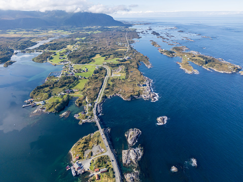 An aerial view of a curving road along a coastal village in Norway. The road links a series of small islands, each with its unique character. Clear surrounding waters display different shades of blue with mountains and clouds in the far background