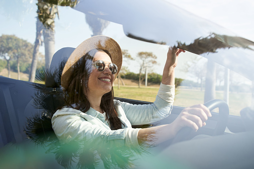 Caucasian young woman in vacation drives a cabriolet smiling in summer. Beautiful and happy and empowered girl having fun in a car. Rent vehicle concept. Lady enjoying the road trip.