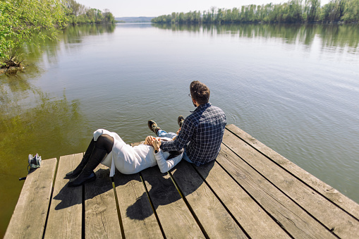 Mid adult interracial couple enjoying their time on a pier by the river or lake. He is sitting, and she is lying with her head in his lap. It's a beautiful sunny day, they look relaxed, happy and in love.
