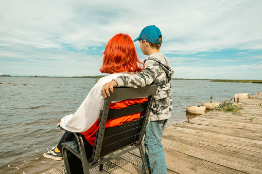 A young girl with red hair, a representative of Generation Z, hugs her younger brother on the shore of the lake, demonstrating family ties and love