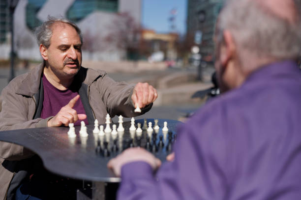 Senior Caucasian plus size man with mustache, concentrating on move chess game. Casual chess match in Gantry Plaza State Park, Long Island City with urban environment background. Looking over the shoulder of opponent