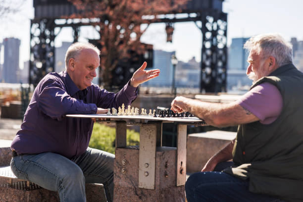 Cheerful senior Caucasian plus-size man raises hand in victory during chess game with a bearded friend at Gantry Plaza State Park table in Long Island City, Queens, New York.