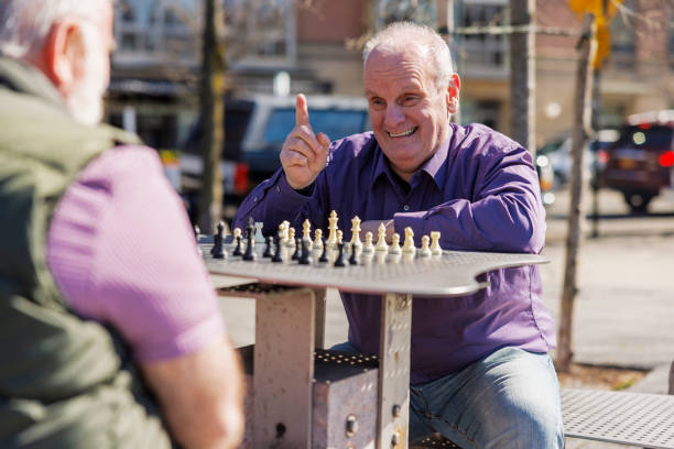 Joyous plus size Caucasian senior man laughing and pointing his finger upwards, while making a winning chess move during game with friend in Gantry Plaza State Park in Long Island City, Queens. Looking over the shoulder of opponent