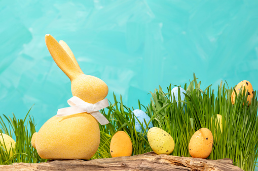 Decorative Easter yellow and blue eggs among fresh green grass and Yellow Easter bunny on old driftwood on turquoise background. Holiday card template with copy space. Horizontal format.