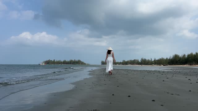 4K Young woman on an empty beach in a white dress walks along the sand holding her shoes, Vietnamese hat on head
