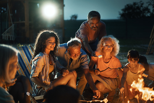 Group of mixed age group of people relaxing by a bonfire and cooking marshmallow candy
